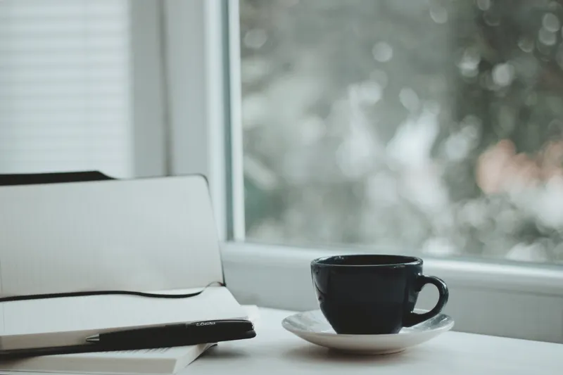 Cup of coffee and a notebook on a table by a bright window